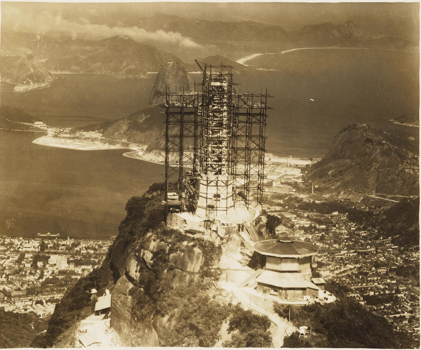 Fotografia antiga do pico do Corcovado com a estátua em construção, envolta em andaimes, e o Pão de Açúcar ao fundo