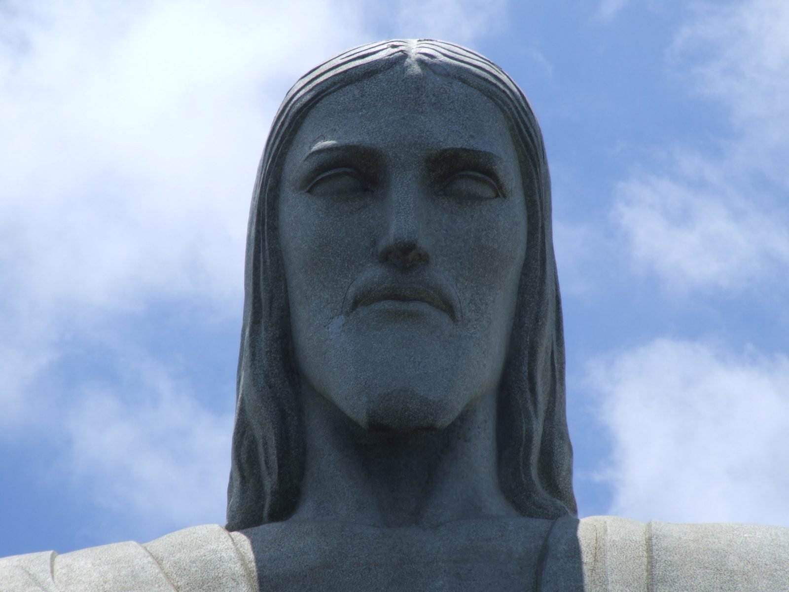 Rosto do Cristo Redentor visto de baixo contra o céu, com a textura do revestimento de pedra-sabão