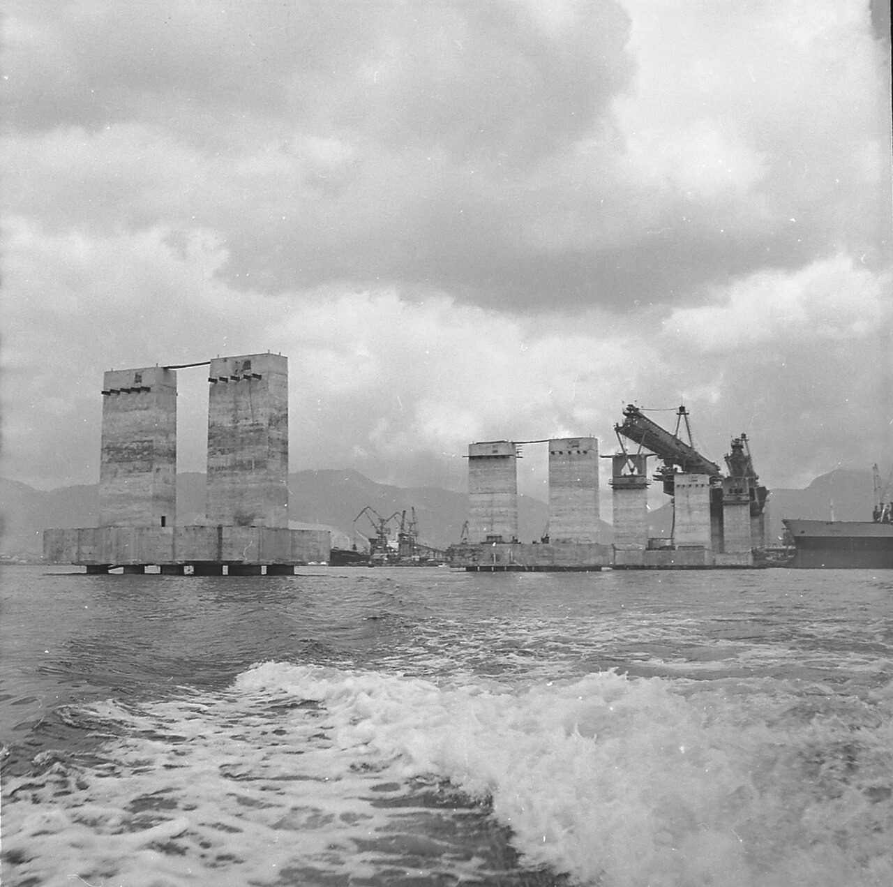Fotografia em preto e branco de pilares da Ponte Rio–Niterói em construção no meio da baía, com guindaste