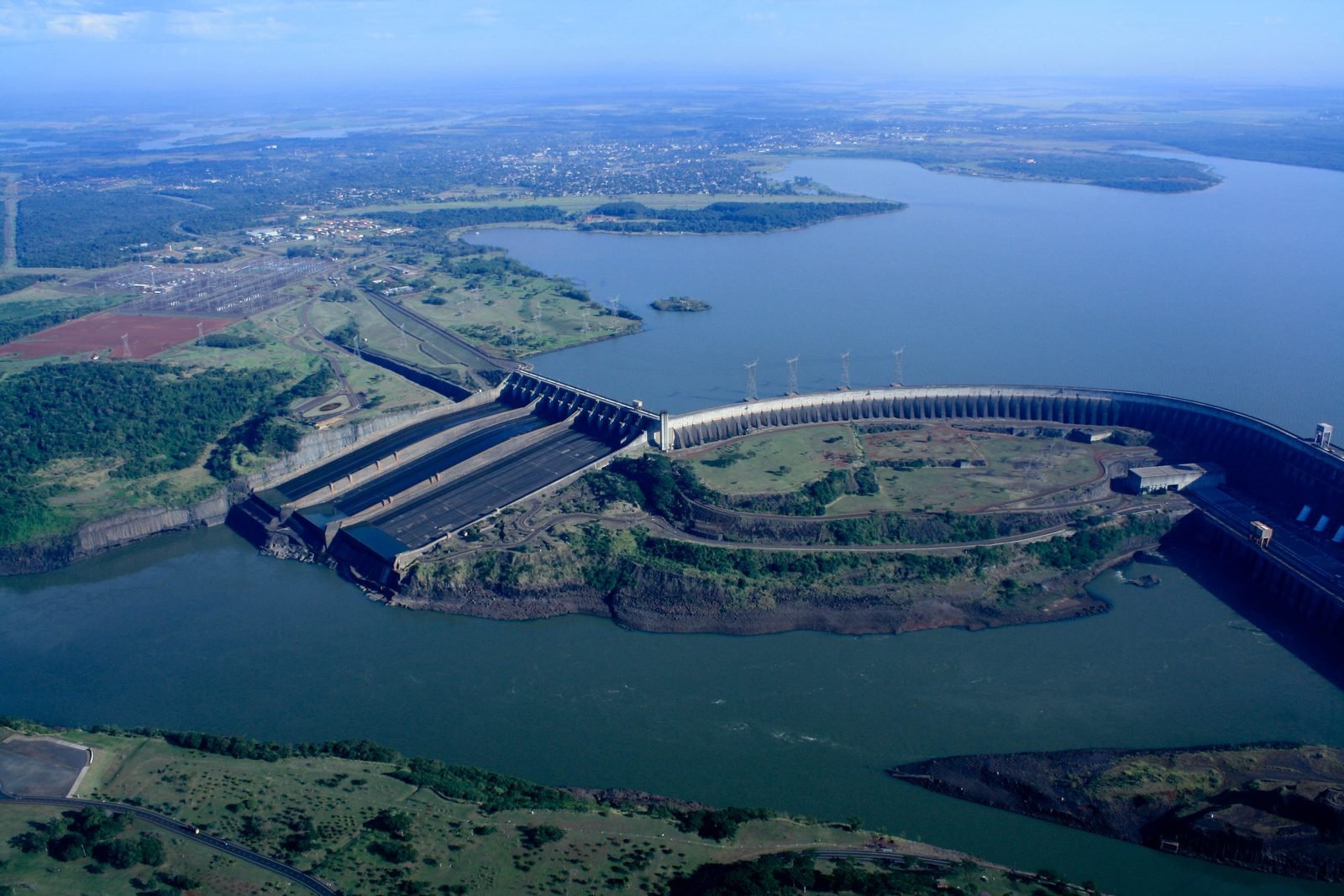 Vista aérea da Usina de Itaipu: o vertedouro à esquerda, a barragem em curva e o reservatório ao fundo, com o rio Paraná em primeiro plano