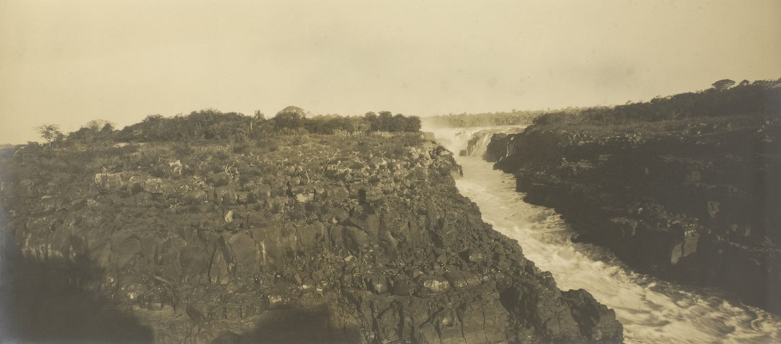 Fotografia antiga em sépia do rio Paraná estrangulado entre paredões de rocha no Salto das Sete Quedas, com quedas d'água ao fundo