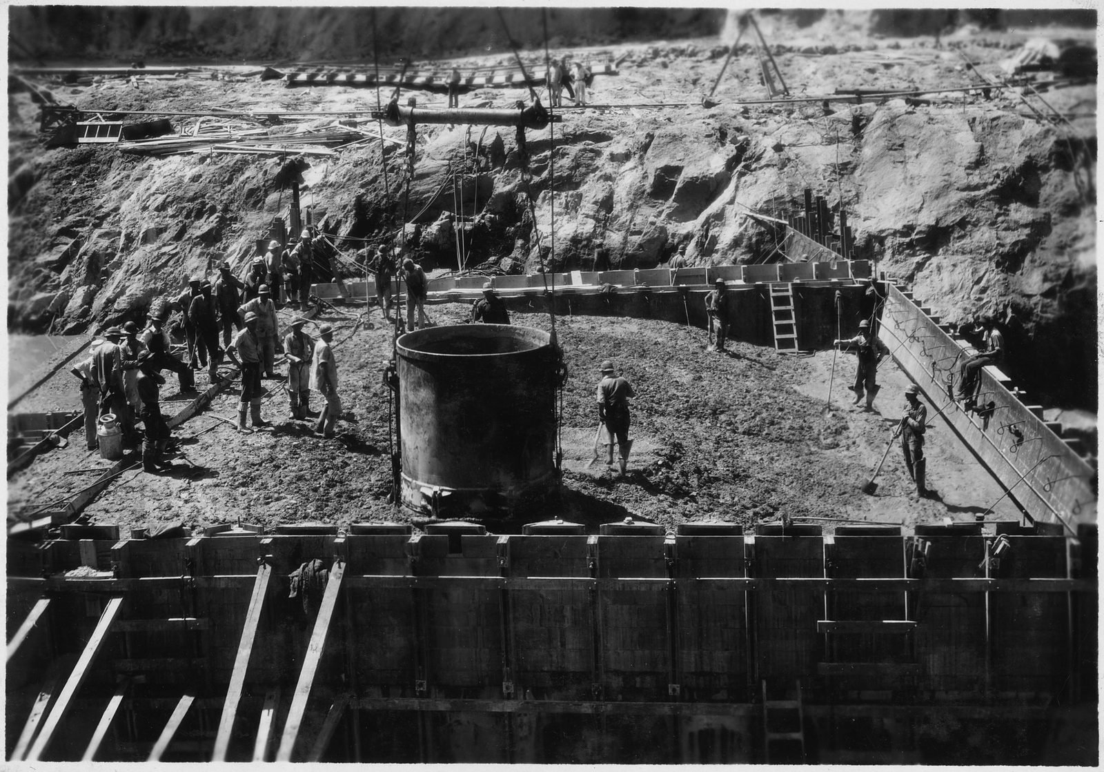 Operários em volta de um grande balde cilíndrico de concreto dentro de uma fôrma de madeira