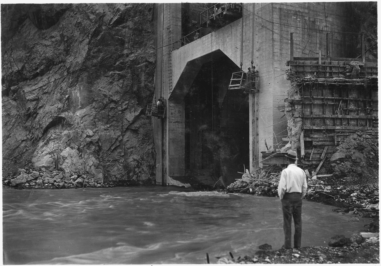 Água do rio saindo por um portal de túnel de concreto no paredão, com um homem de camisa branca olhando de perto