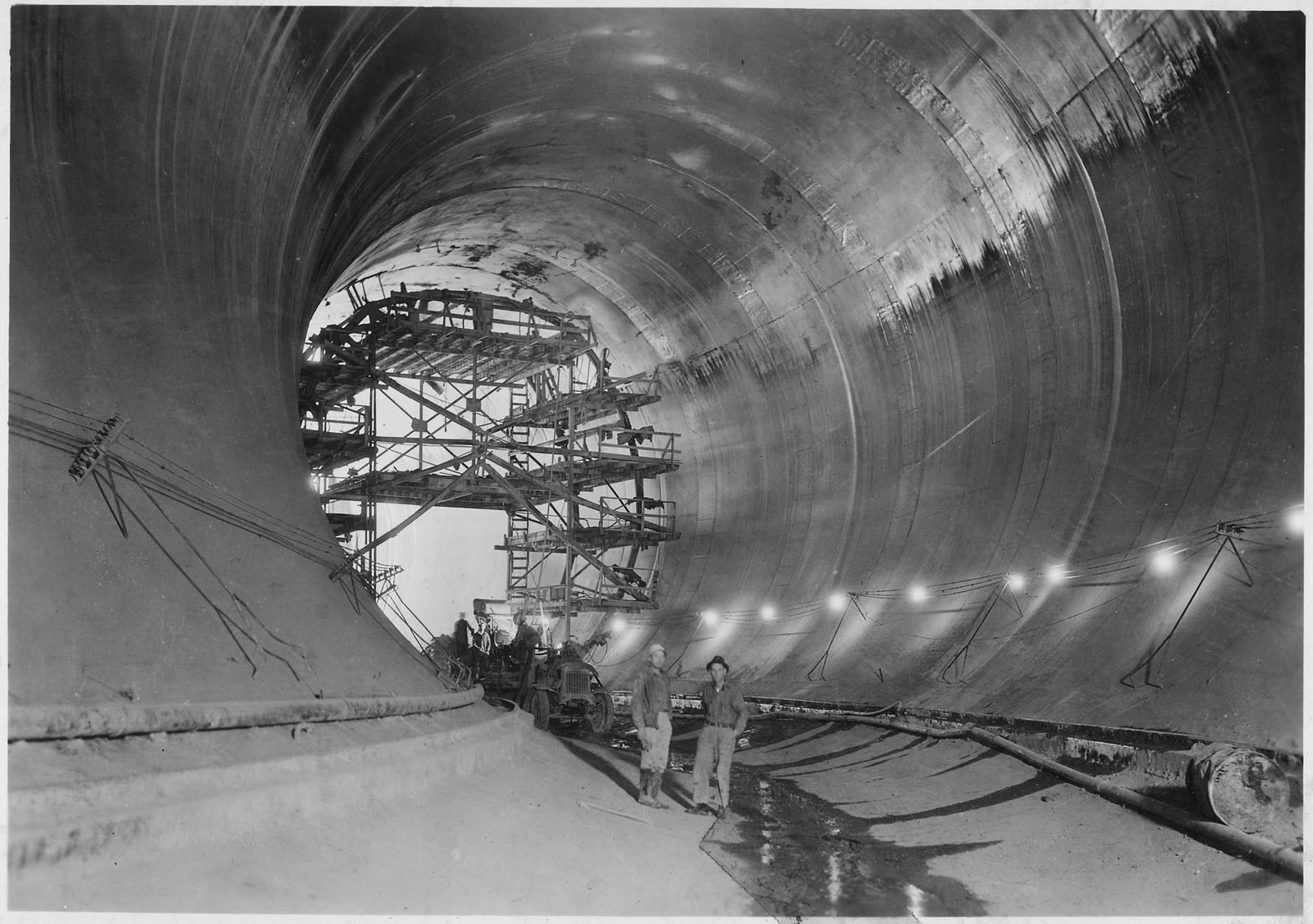 Interior de um túnel de desvio já revestido de concreto, com andaime metálico e dois homens no piso