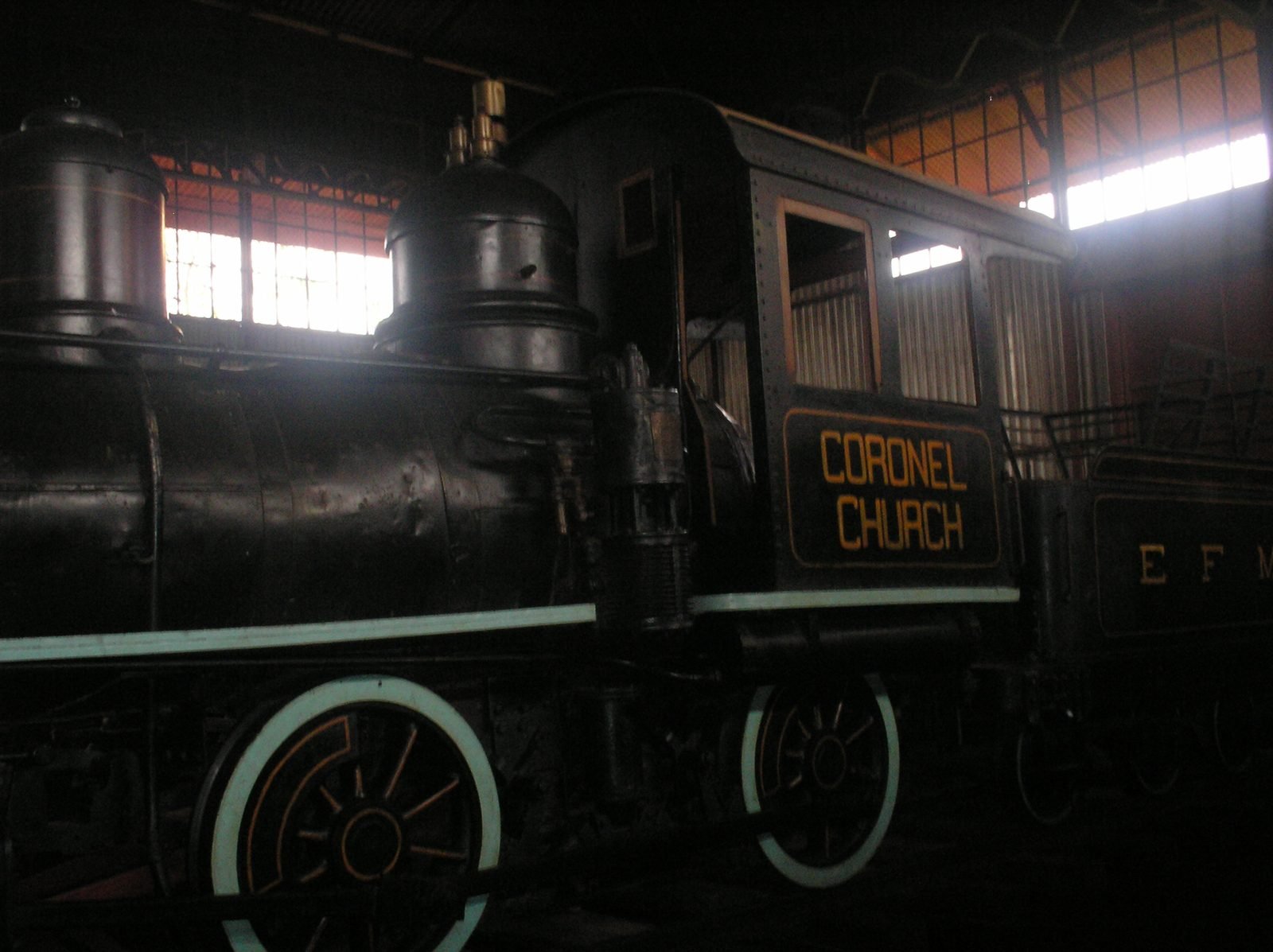 Locomotiva a vapor preta com rodas de aro claro e a inscrição Coronel Church na cabine, dentro de um galpão