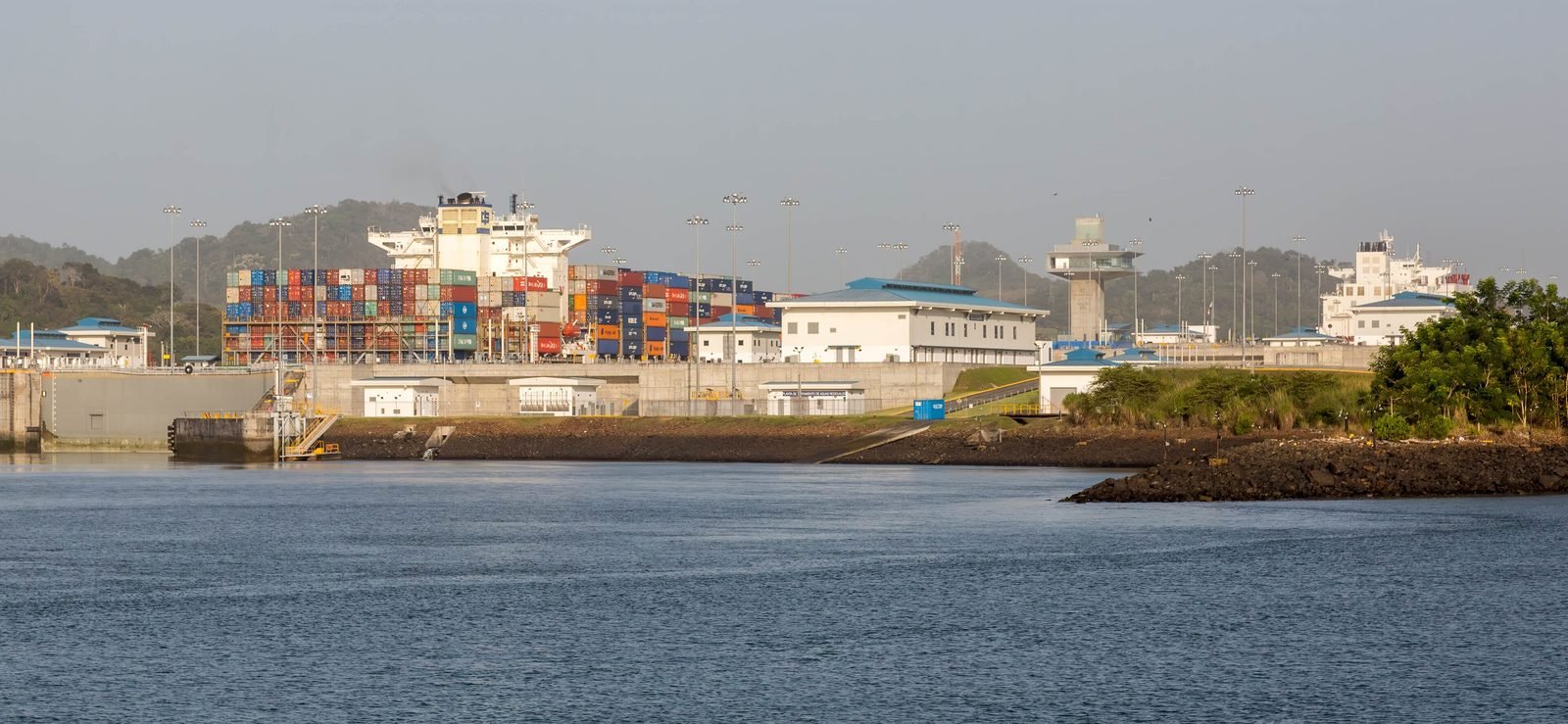 Navio porta-contêineres muito carregado dentro das eclusas novas, atrás de edifícios brancos e de uma torre de controle, com água em primeiro plano