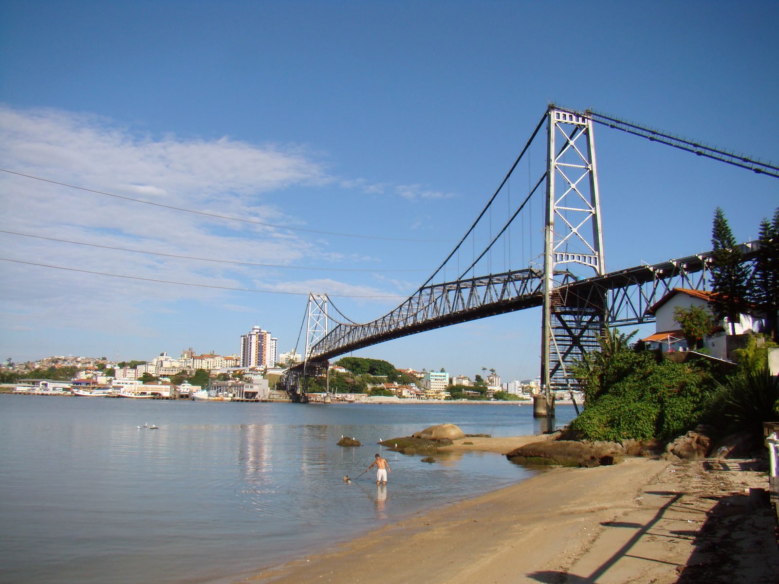 Ponte pênsil de aço cinza sobre água calma, com uma torre treliçada alta à direita, a corrente descendo em curva até a treliça do vão central e uma praia com pedras em primeiro plano