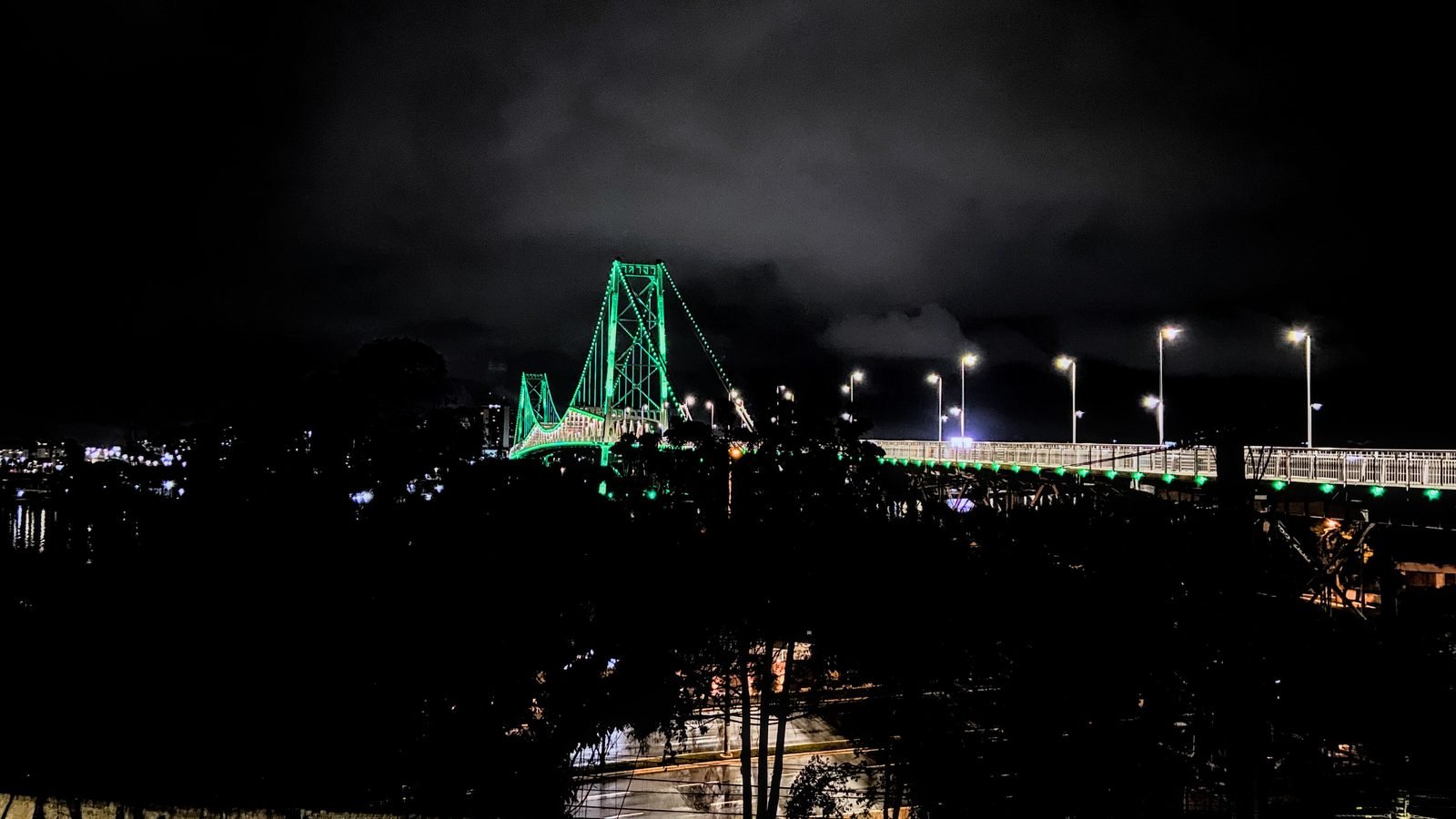 A ponte à noite com as torres e a corrente contornadas por luzes verdes, postes acesos sobre a pista e a cidade escura ao redor