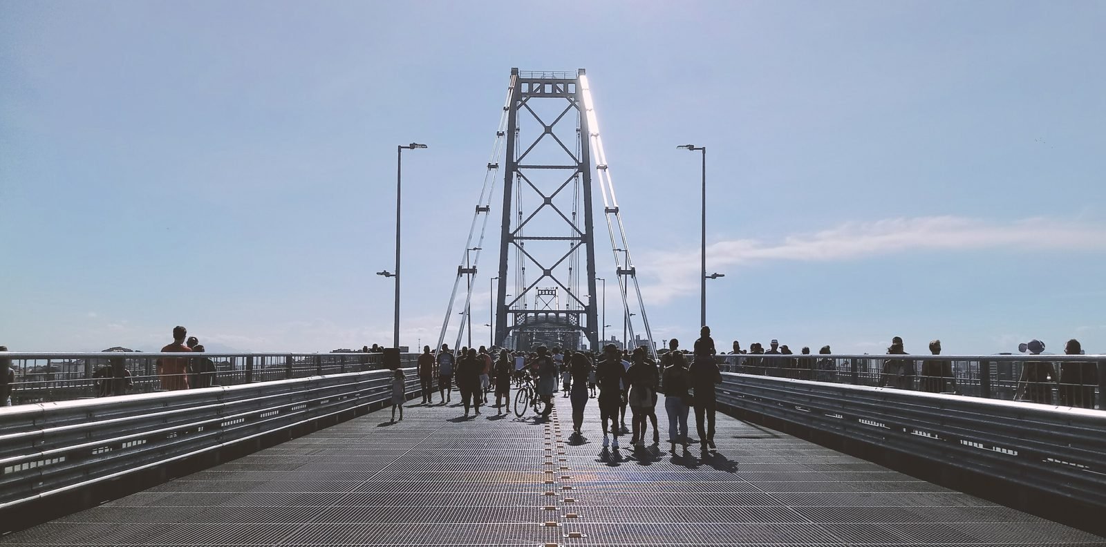 Pessoas caminhando sobre o piso de grade metálica da ponte em direção a uma torre treliçada, sob céu claro
